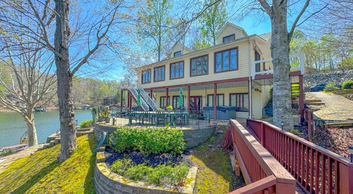 The exterior of the lakefront home is shown, featuring multiple levels with large windows. A deck with seating faces the lake, surrounded by landscaped greenery. The tranquil water and trees are visible in the background, creating a serene outdoor setting.