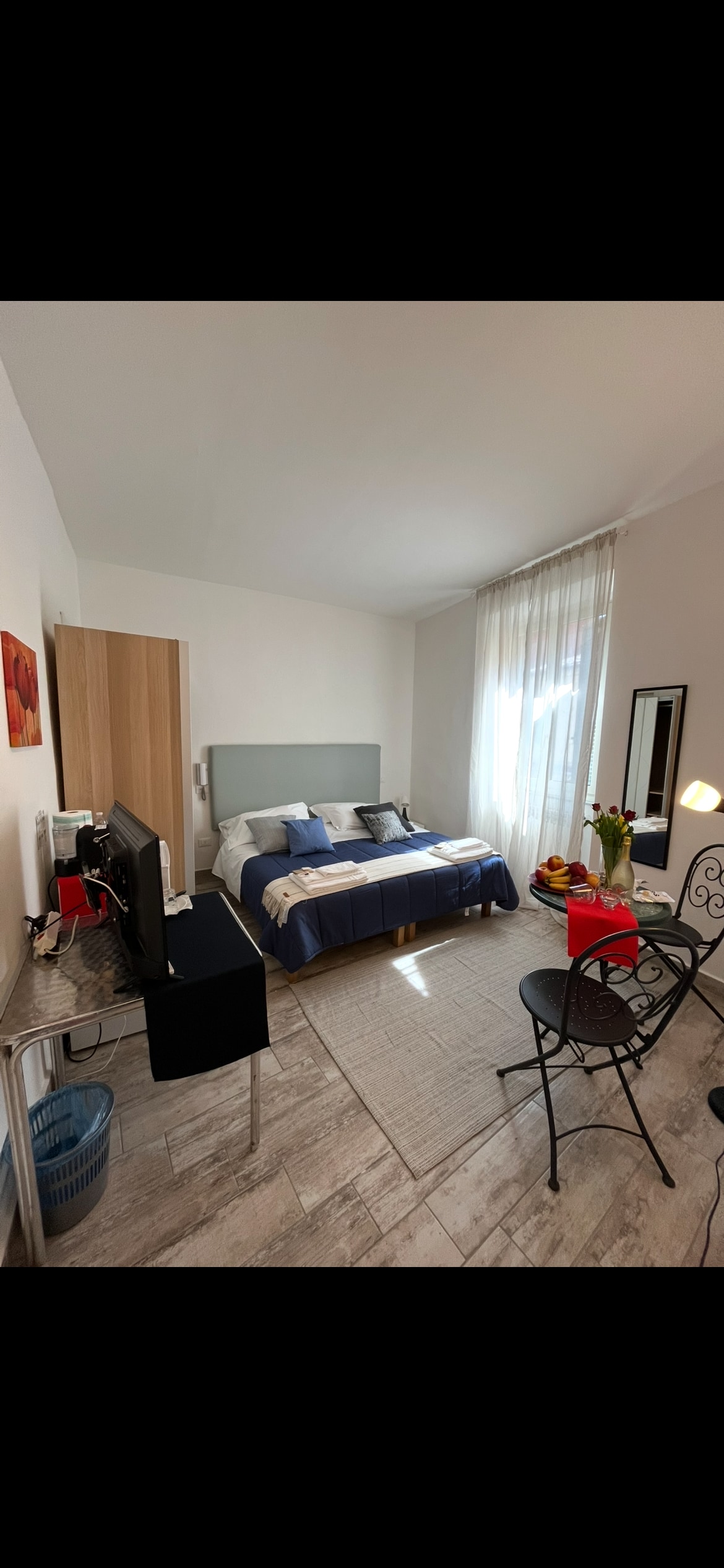 A comfortable sleeping area is furnished with a spacious bed featuring blue and white linens. A small desk with a television is positioned adjacent to the bed. Natural light enters through large curtains, illuminating a cozy seating area with a small table and two chairs.