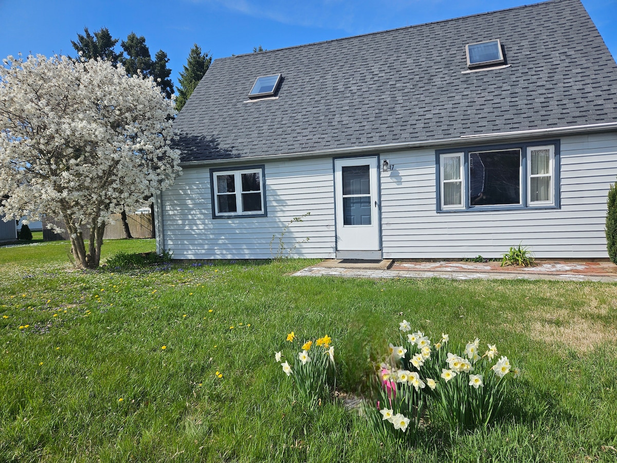 The exterior of a charming two-story house is shown, featuring a gray roof and white siding. A flowering tree stands to the left, while colorful daffodils bloom in the front yard. Natural light is captured in the windows, enhancing the inviting appearance of the entrance.