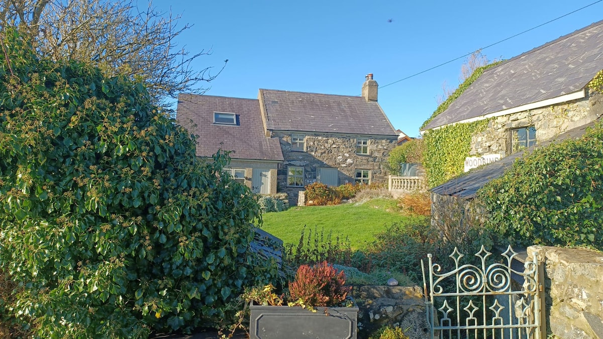 A stone cottage surrounded by lush greenery is seen against a clear blue sky. The structure features a slate roof and charming stone walls. A well-maintained garden and patio area are visible, with various plants and shrubs adding to the peaceful outdoor setting.