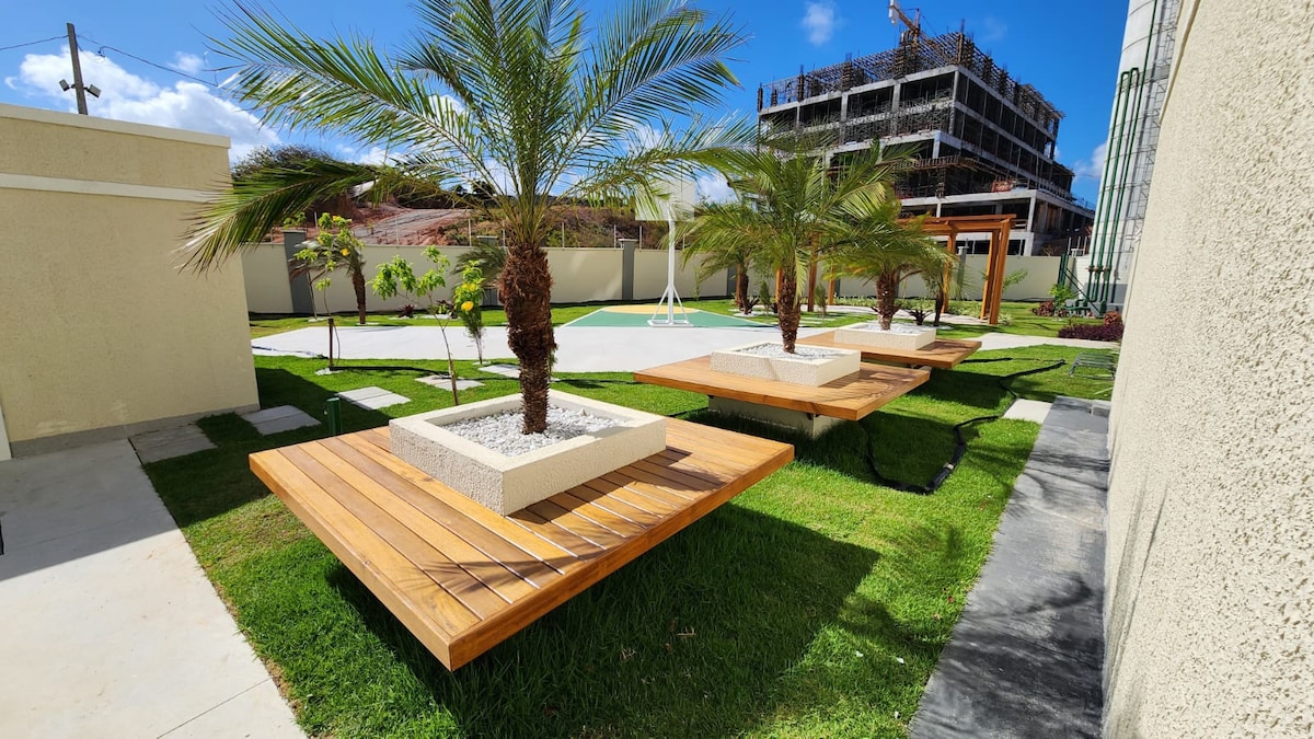 An outdoor area features landscaped green spaces with neatly arranged wooden platforms surrounded by palm trees. The setting incorporates clean lines and modern design, with a spacious pool visible in the background, creating an inviting space for relaxation.