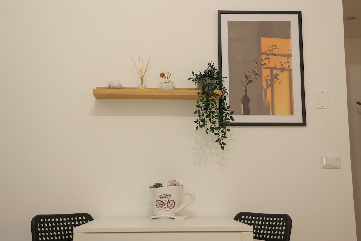 A neat dining area features a minimalist white table with two black chairs. A decorative shelf above holds various items including a small vase and green plant. A framed art piece adds a splash of color to the wall, enhancing the room's ambiance.