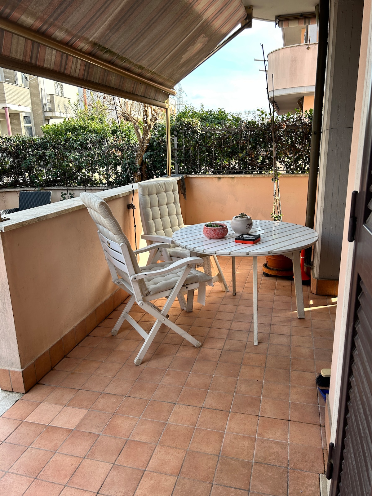 A private outdoor space is captured, featuring a round table surrounded by two chairs. A potted plant is visible, along with a teapot and small decorative items on the table. The area is sheltered by an awning, and a lush green hedge provides privacy.