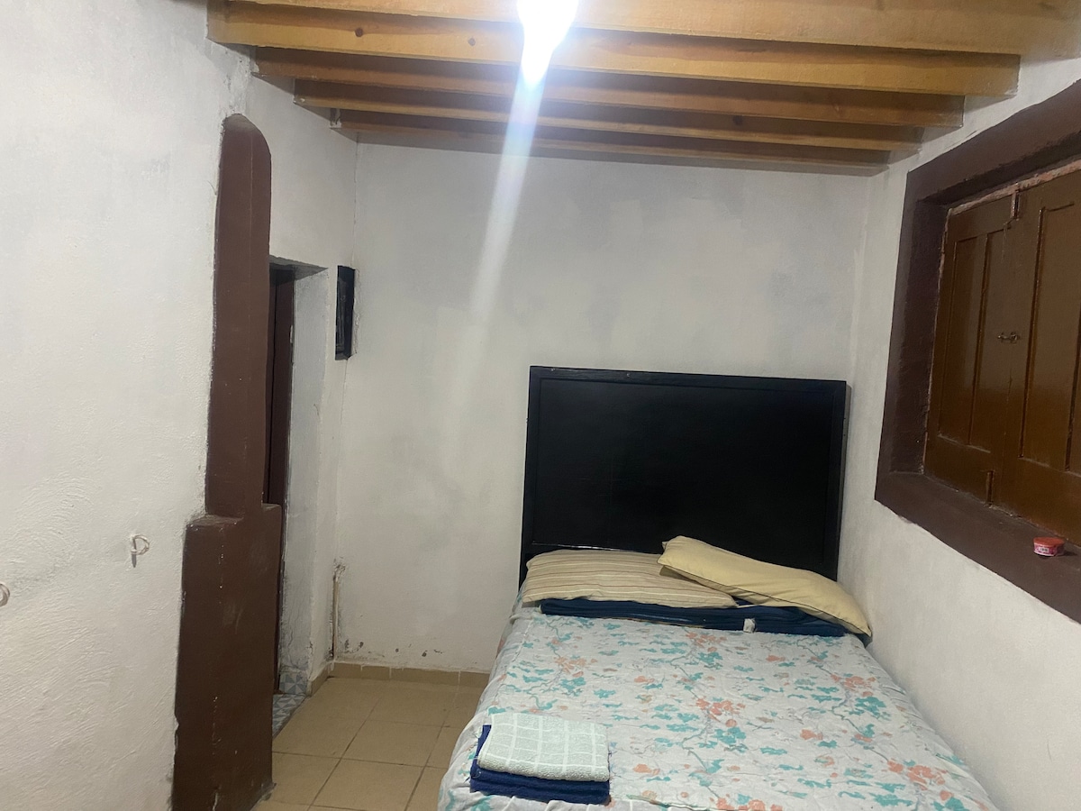 A simple sleeping area is presented with a spacious bed dressed in a floral-patterned blanket. Light-colored pillows are arranged at the head of the bed. Exposed wooden beams run across the ceiling, and a window with wooden shutters permits natural light to enter the room.