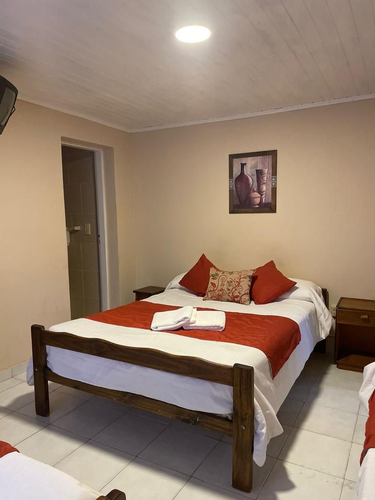 A room is featured with a spacious bed dressed in white linens and accented with orange pillows. A small wooden nightstand is positioned next to the bed. A piece of artwork adorns the wall, and a doorway leads to a bathroom.