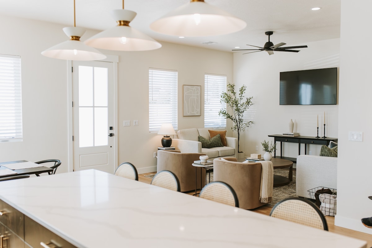 A bright and inviting living space features comfortable seating arrangements with neutral tones. Large windows allow natural light to fill the room. A modern entertainment unit holds a flat-screen television. The kitchen area showcases a sleek counter, blending functionality with style.
