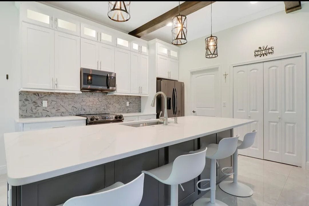 A modern kitchen features sleek cabinetry and an island with a white countertop. Three high-backed stools are positioned along one side. Pendant lights hang from the ceiling, casting soft illumination, while stainless steel appliances offer a contemporary touch.