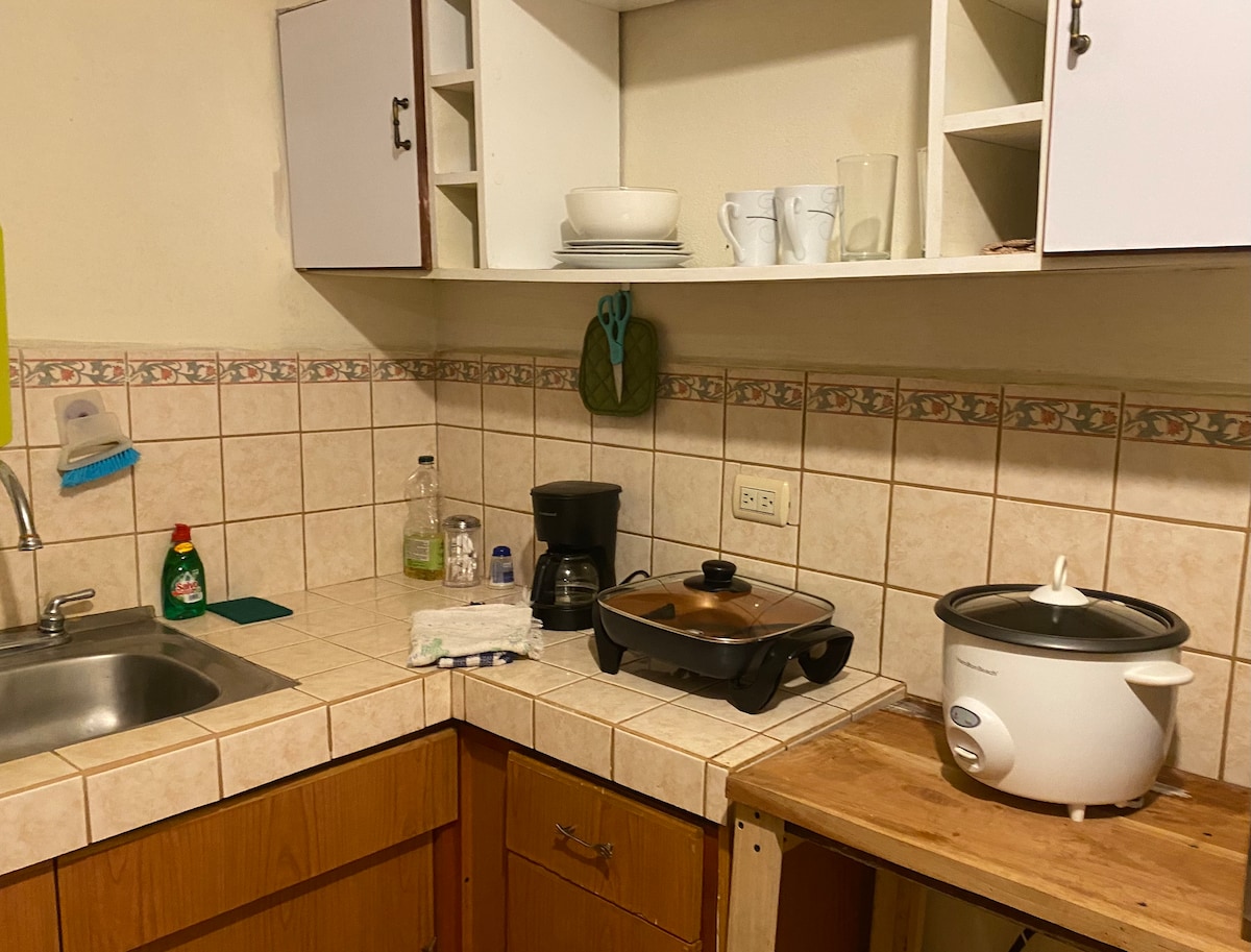 A functional kitchen area is displayed, featuring a sink, countertop, and cabinets. A coffee maker and cooking utensils are positioned on the counter, alongside a rice cooker and a frying pan. Glassware is neatly organized on the shelves above, contributing to the kitchen's practicality.