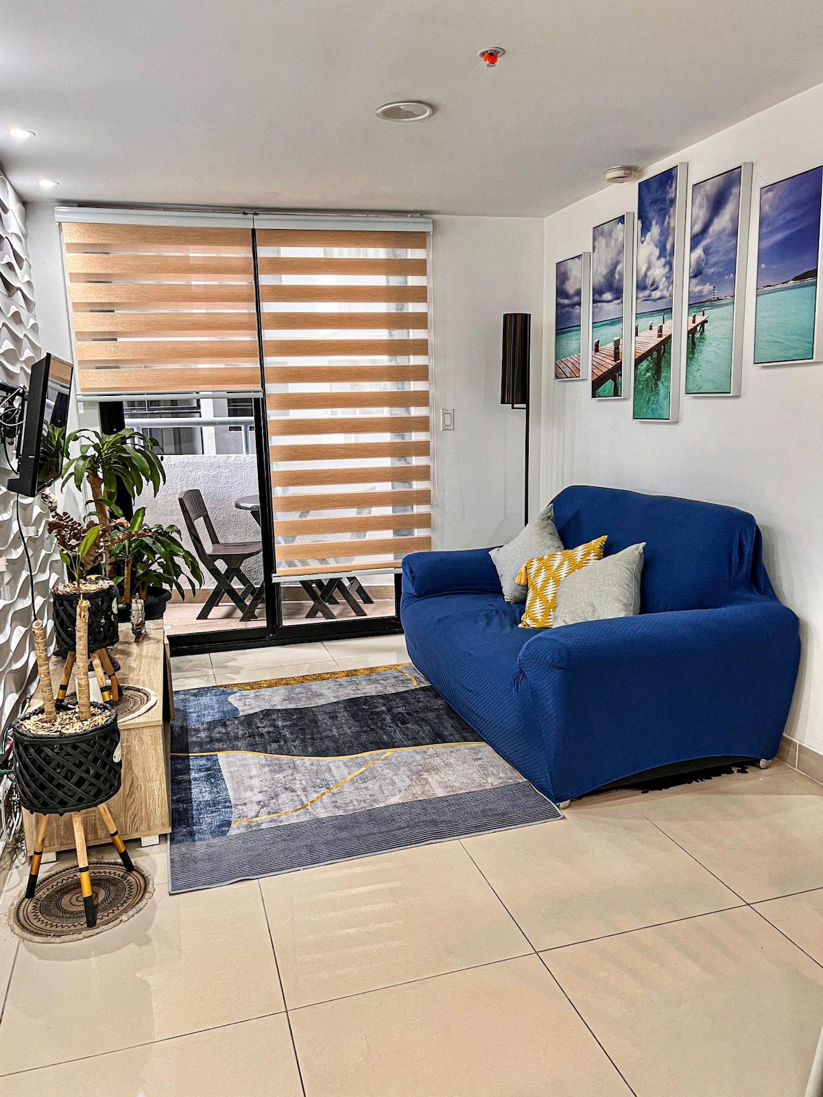 A living area features a blue sofa adorned with decorative cushions, positioned next to an indoor plant. Light-colored tiles reflect natural light from large sliding doors, which lead to a small outdoor sitting area. Decorative wall art complements the stylish design.