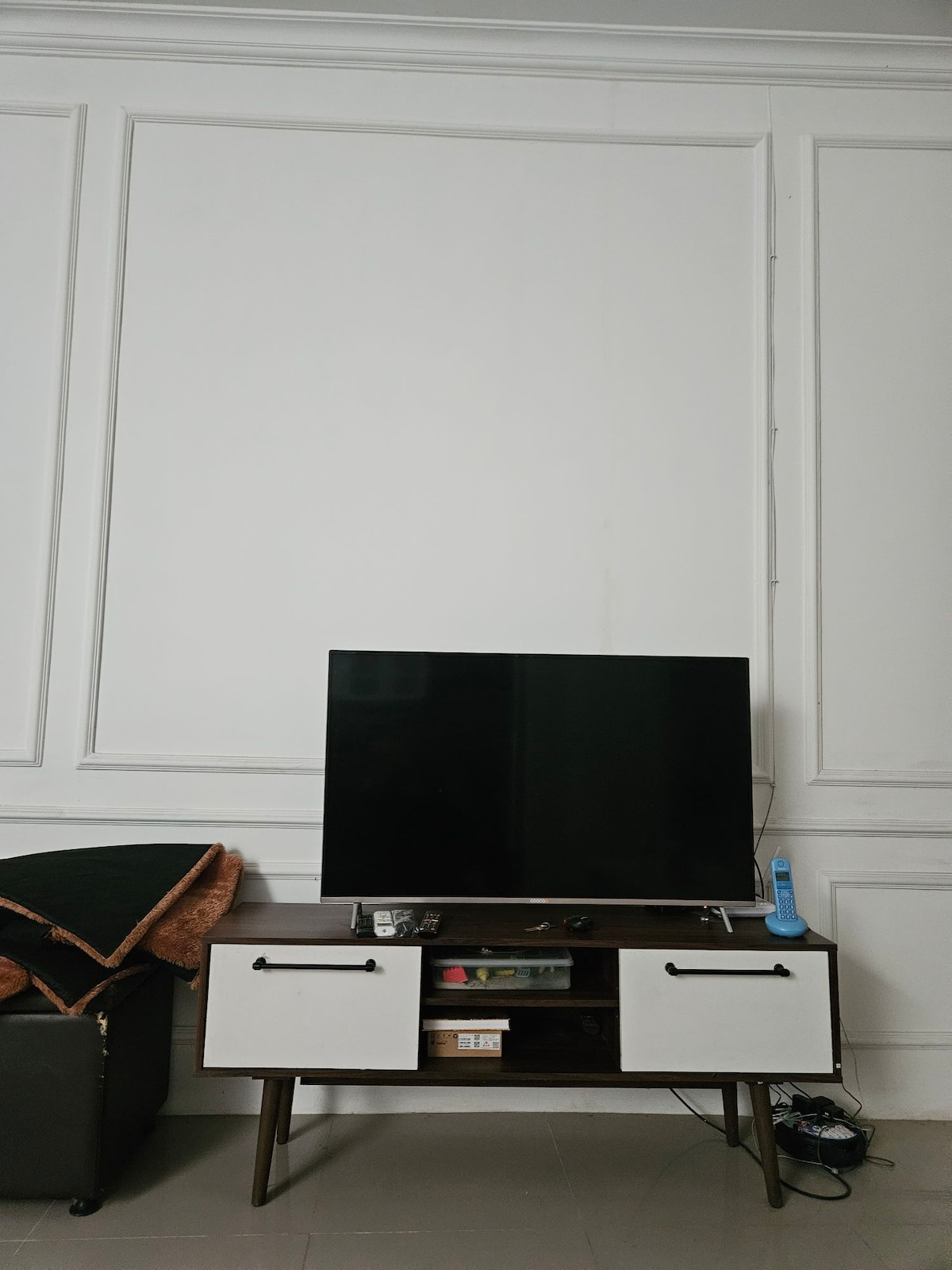 A flat-screen television is mounted on a sleek cabinet with two white drawers. The wall behind features decorative paneling, while a dark sofa with an orange throw blanket is positioned nearby, adding a touch of color to the neutral space.