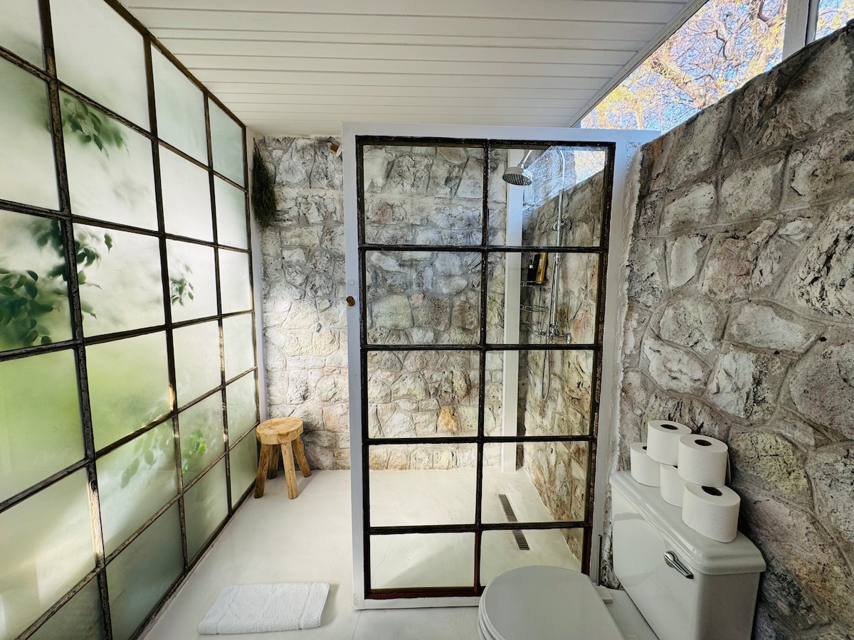 A modern bathroom is presented with stone walls featuring a natural texture. A glass shower enclosure offers a clear view of the shower area. A wooden stool sits beside the shower. The space contains essential toiletries and is illuminated by natural light entering through a window.