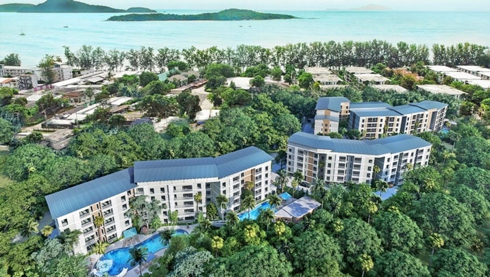 An aerial view of the condominium complex set amidst lush greenery, displaying multiple buildings surrounded by tropical landscaping. The outdoor swimming pools and the nearby beach are visible, along with the serene blue sea and islands in the distance, creating a tranquil setting.