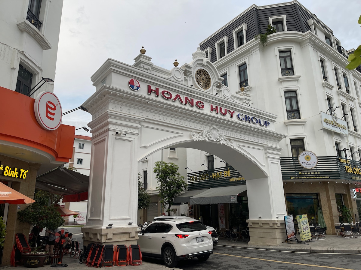 An elegant archway featuring the name 'Hoang Huy Group' prominently displayed above. The structure is adorned with a clock and is surrounded by multiple buildings. A white vehicle is parked beneath the arch, while outdoor seating can be seen nearby.