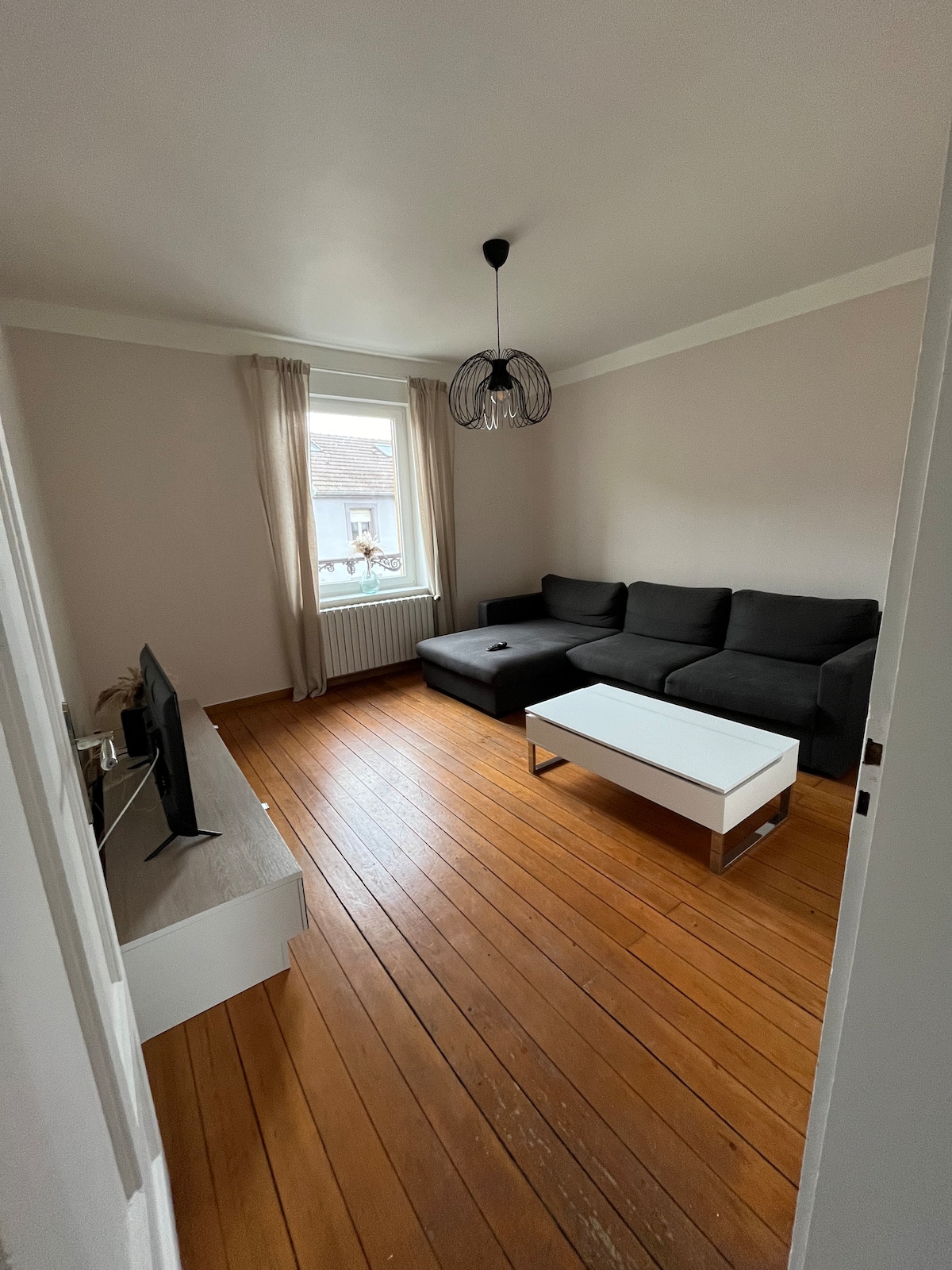 A welcoming living room features a sleek black sofa and a minimalist white coffee table. Natural light enters through a window, complemented by light curtains. A modern television is set against the wall, enhancing the room’s functionality in a relaxed setting.