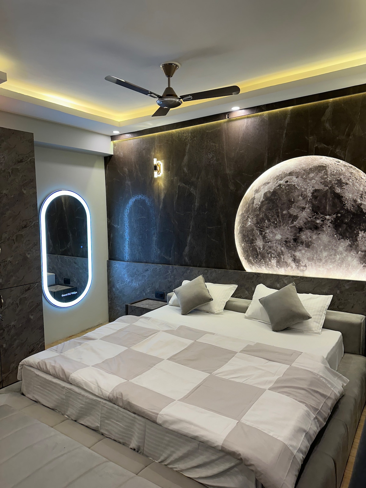 A serene sleeping area features a spacious bed with a light-colored quilt, complemented by matching gray cushions. A large moon-shaped wall art piece creates a focal point, while subtle lighting enhances the room’s calming ambiance. A stylish mirror adds a functional element.