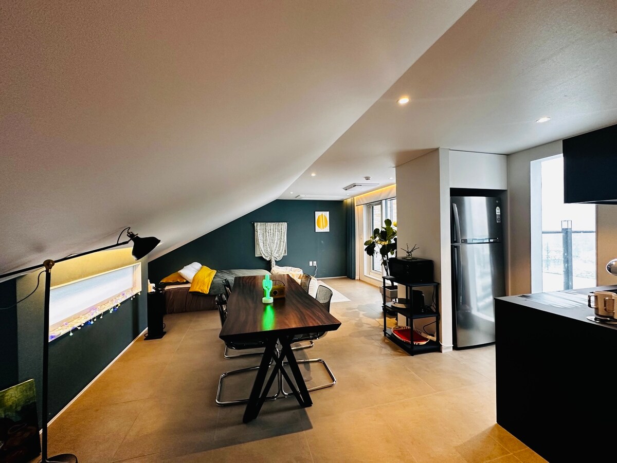 An open living space features a large wooden table set for dining and a comfortable seating area with a sofa. Large windows allow natural light to fill the room, while modern appliances are present in the kitchen area. A projector screen is visible, ready for entertainment.