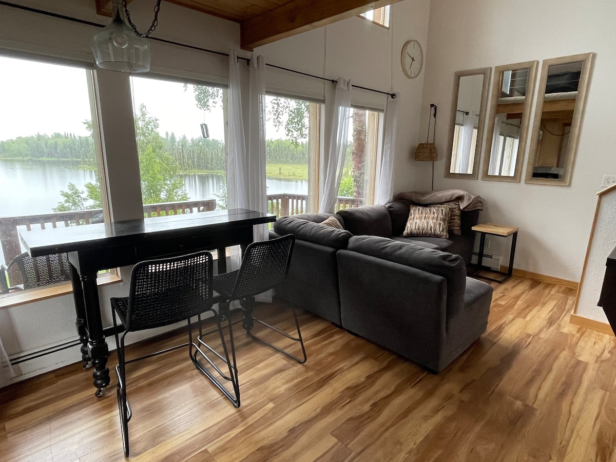 A cozy living area features a sectional couch and a small dining table with four chairs. Large windows provide views of the lake and surrounding greenery. Soft natural light brightens the wood floors and walls, creating a serene atmosphere.