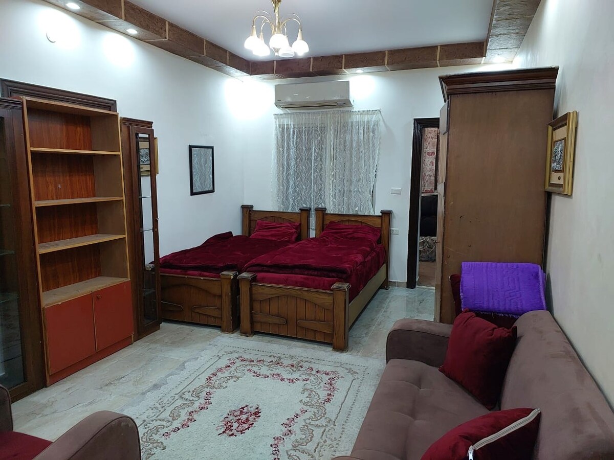 A comfortable room features two spacious beds dressed in rich red bedding. Nearby, a cozy sofa offers additional seating. Wooden shelving and a cabinet provide storage, while a sheer curtain allows natural light to enter through the window.