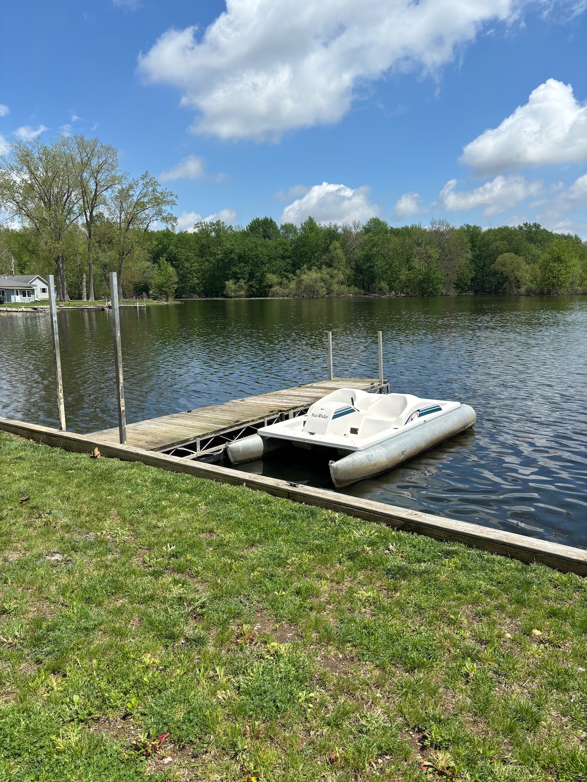 A peaceful lake scene is captured, showcasing a wooden dock extending over calm waters. A paddleboat is moored alongside the dock, surrounded by lush greenery and trees lining the shore. Bright clouds float in the blue sky above, enhancing the tranquil atmosphere.