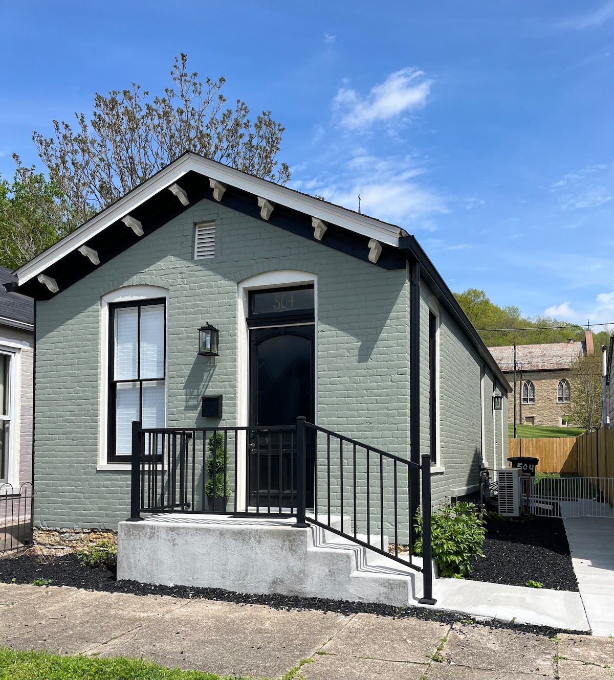 The exterior of a cozy cottage is showcased, featuring a light green brick facade. A short set of stairs leads up to the front door, complemented by a black railing. The surrounding area includes black mulch and shrubs, with blue skies visible above.