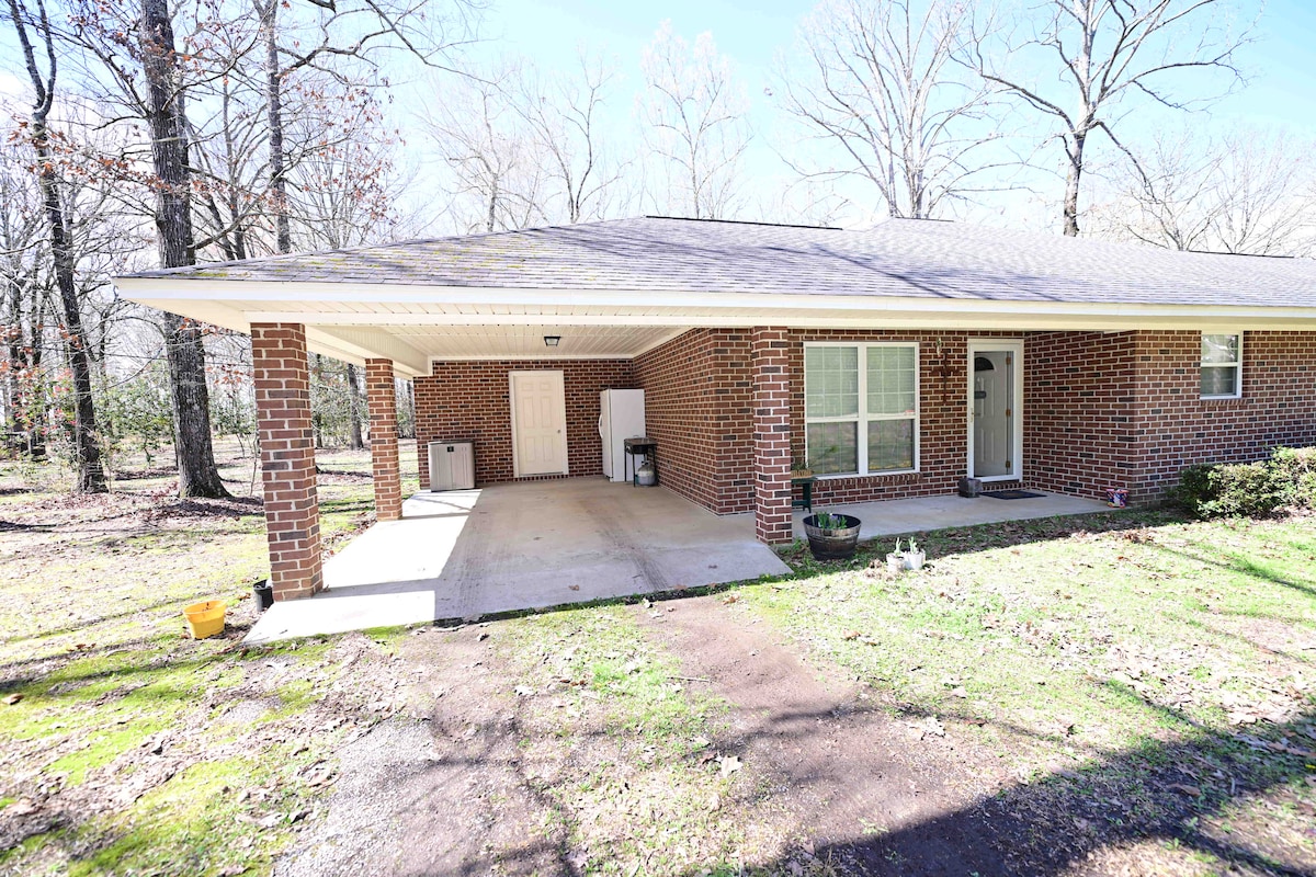 A spacious exterior view of a brick home is presented, featuring a covered carport and large window. Surrounding greenery includes patches of grass and trees, providing a tranquil setting. A storage area is visible in the carport, enhancing functional outdoor space.