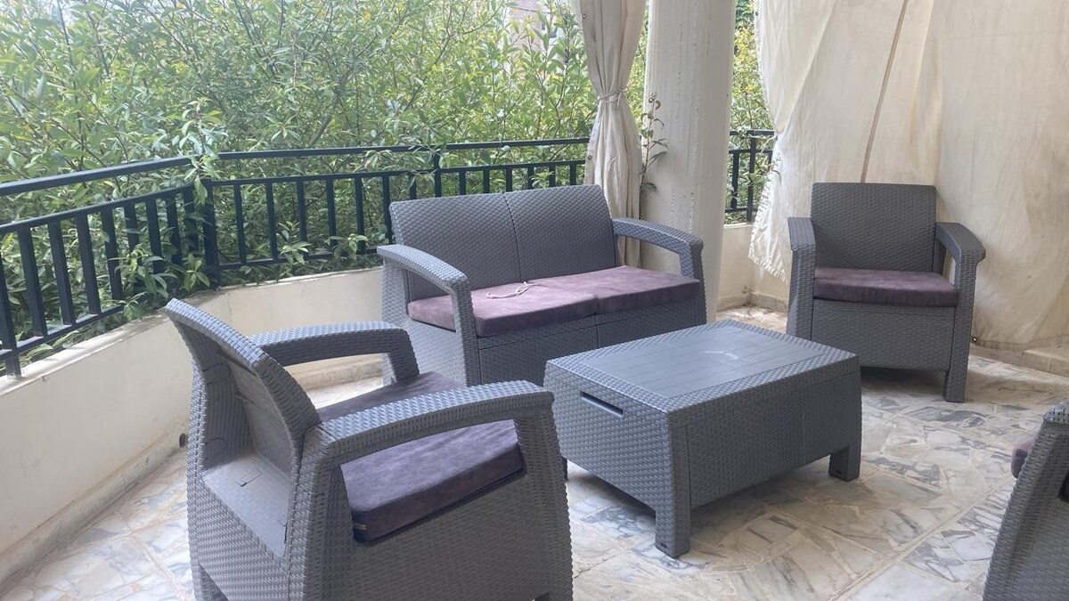 An outdoor seating area is presented, featuring a comfortable sofa and two chairs, all crafted from gray wicker. A coffee table is positioned centrally, providing an inviting space for relaxation. Lush greenery can be seen through a railing, enhancing the serene environment.