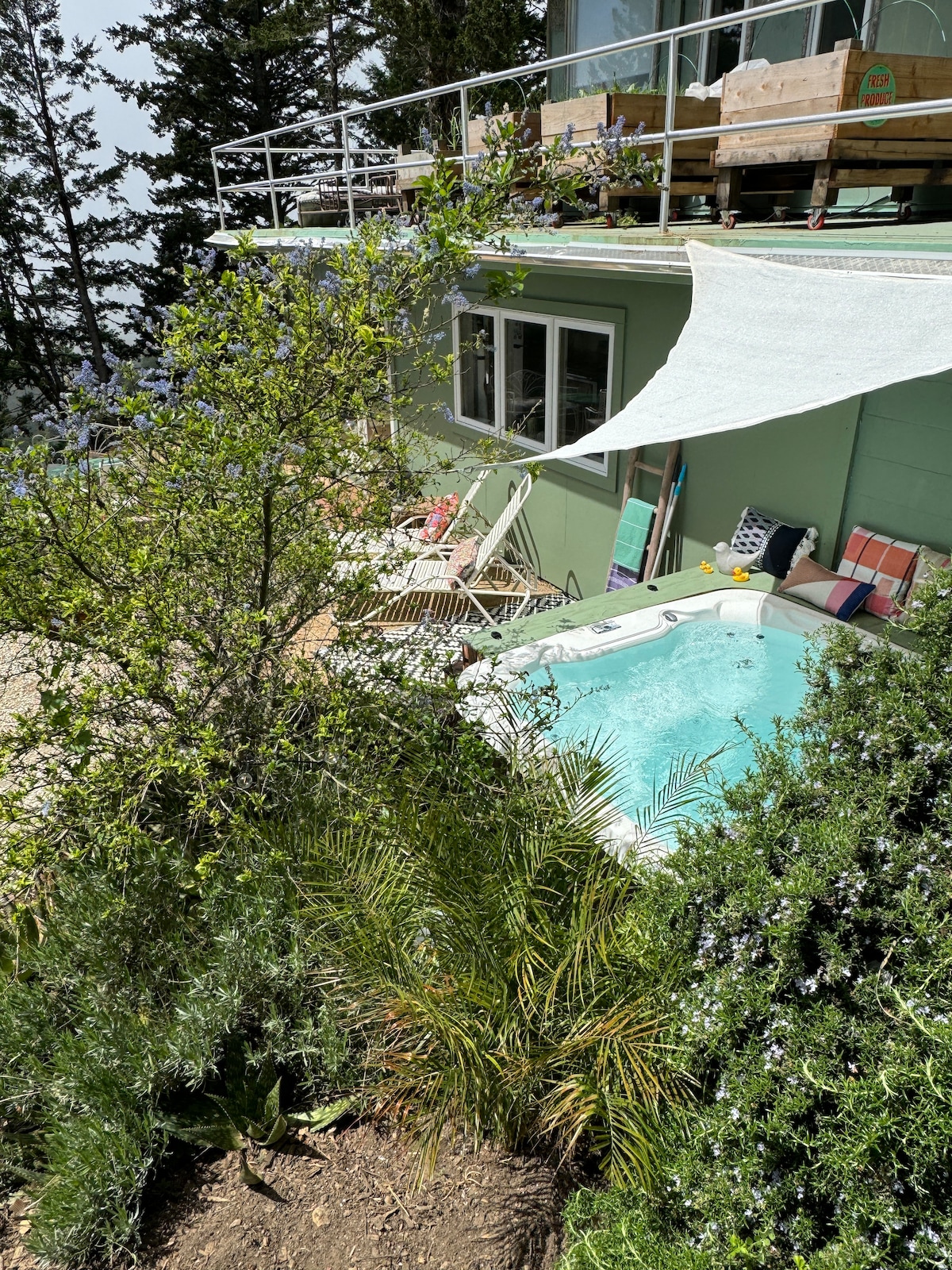 A secluded outdoor area features a hot tub surrounded by lush greenery. A shaded patio sits nearby with white lounge chairs, while a portion of the building is visible in the background, incorporating large windows and a deck area adorned with outdoor furnishings.