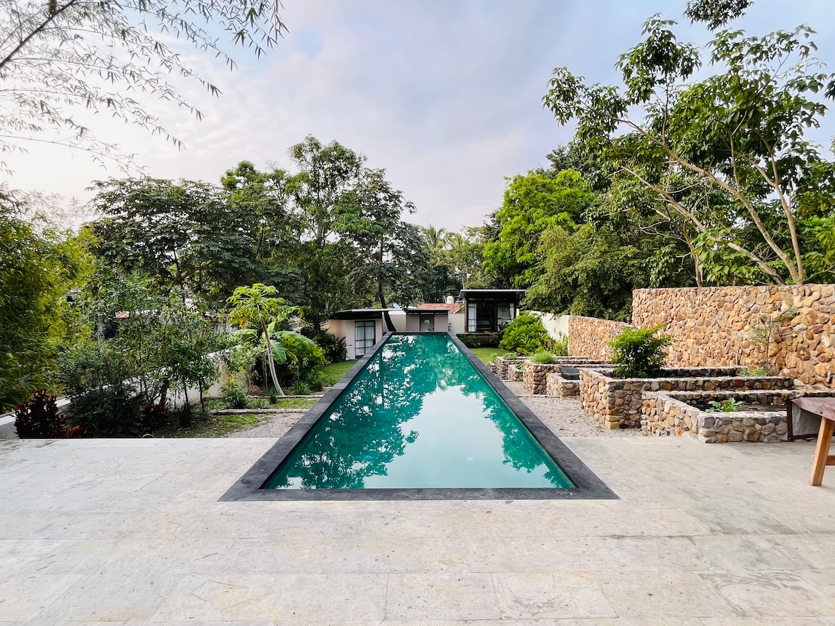 An elongated salt-water pool reflects the surrounding greenery, framed by natural stone walls and mature trees. The pool area features smooth stone flooring, providing a seamless transition to the lush garden scenery. The tranquil environment invites relaxation and connection with nature.