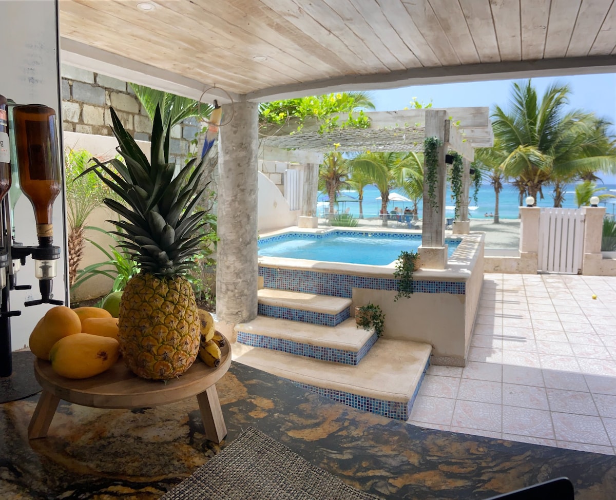 A bright and airy space featuring a view of the outdoor pool surrounded by tropical greenery. A table with fresh fruits, including a pineapple and mangoes, is positioned on the countertop. Sunlight illuminates the area, enhancing the connection between indoor and outdoor settings.