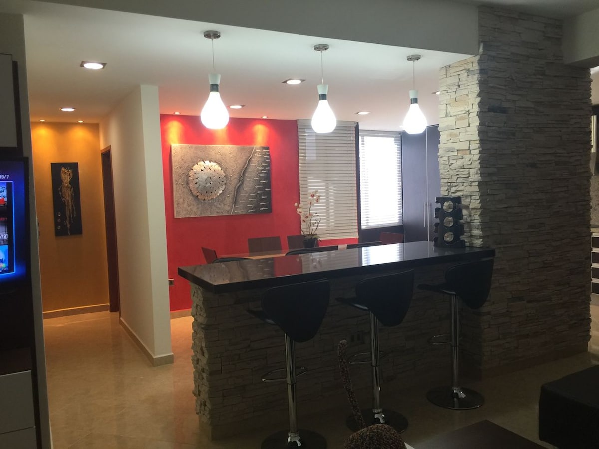 An open-concept living space features a kitchen island with three black bar stools. The area is highlighted by a mix of warm and cool wall colors, and decorative wall art enhances the ambiance. Soft lighting from pendant fixtures illuminates the room, creating a welcoming environment.