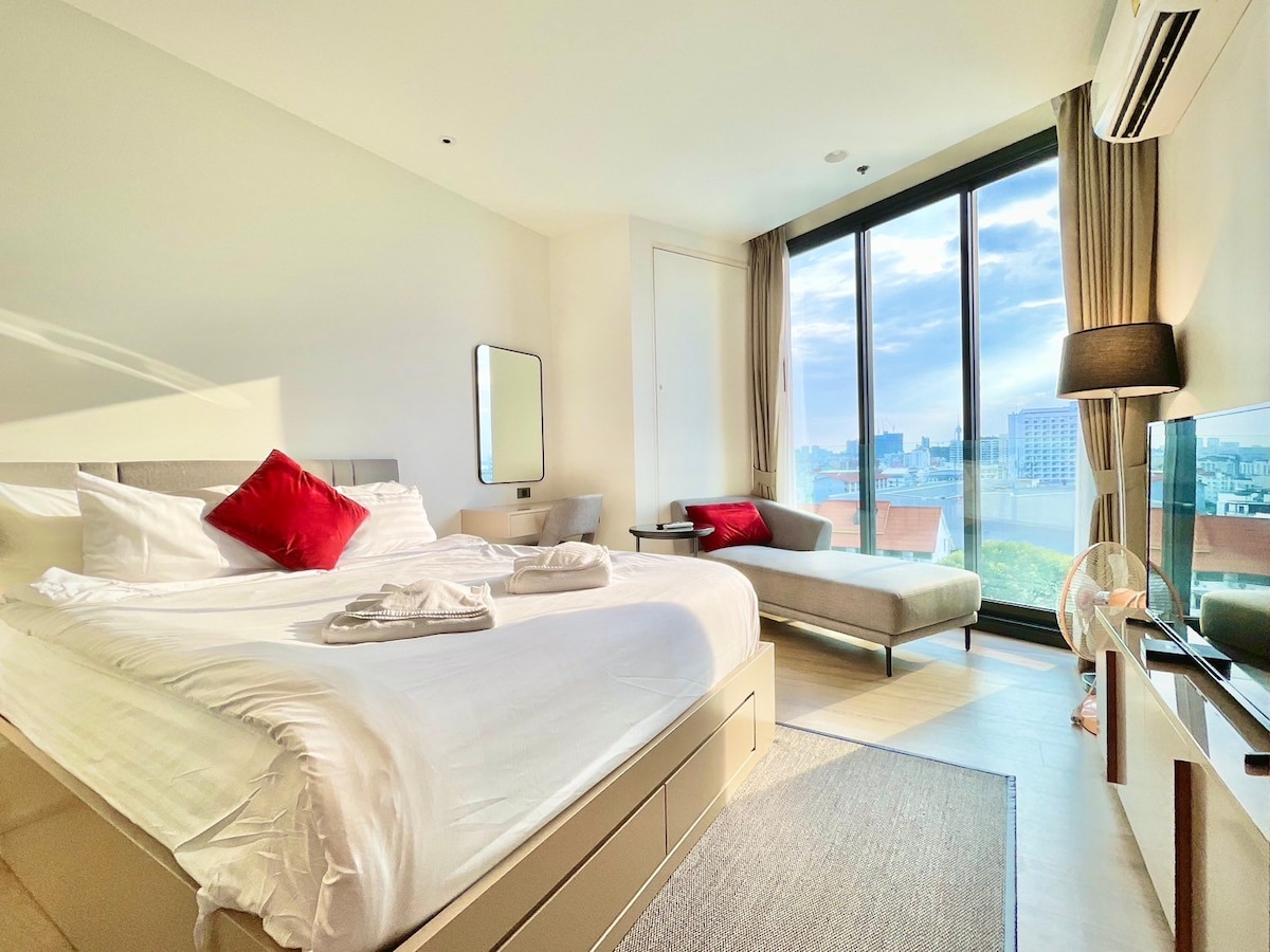 A modern room features a spacious bed adorned with crisp linens and decorative pillows. A large window offers expansive city views, allowing natural light to illuminate the space. A cozy daybed is positioned beside the window, complemented by a minimalist design.