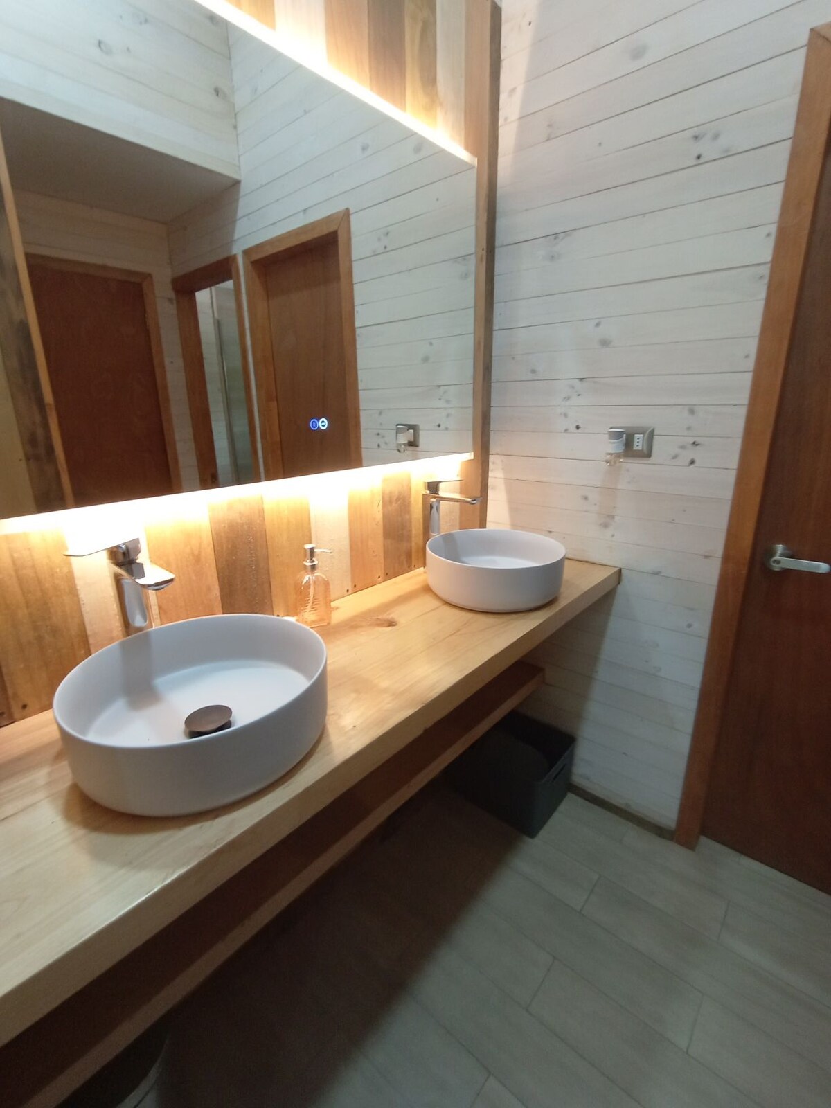 The bathroom features a wooden countertop with two circular sinks and modern faucets prominently displayed. A well-lit mirror extends above the sinks, reflecting light across the space. Warm wood paneling creates an inviting backdrop, while neutral tiles add a clean finish to the floor.
