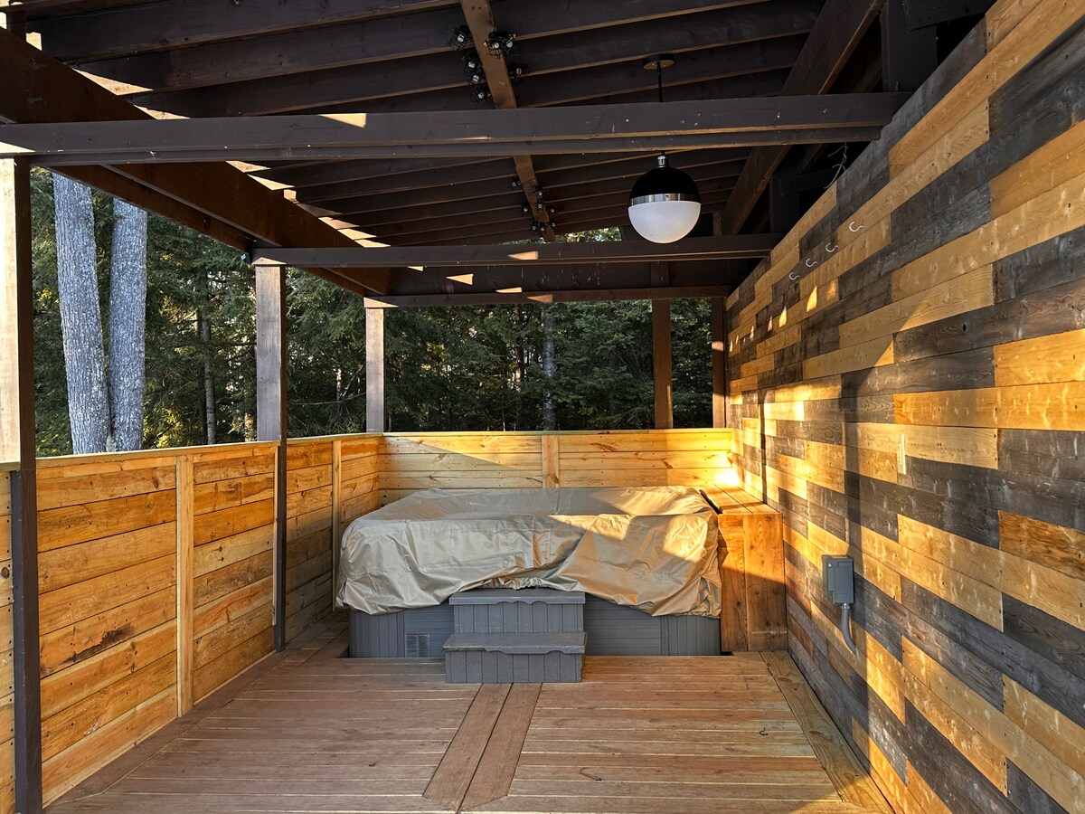 A covered outdoor area features wooden walls and a spacious deck. A hot tub, covered with a protective tarp, is situated in the corner. Natural light filters through the structure, highlighting the wood grain and warm tones of the surrounding decor.