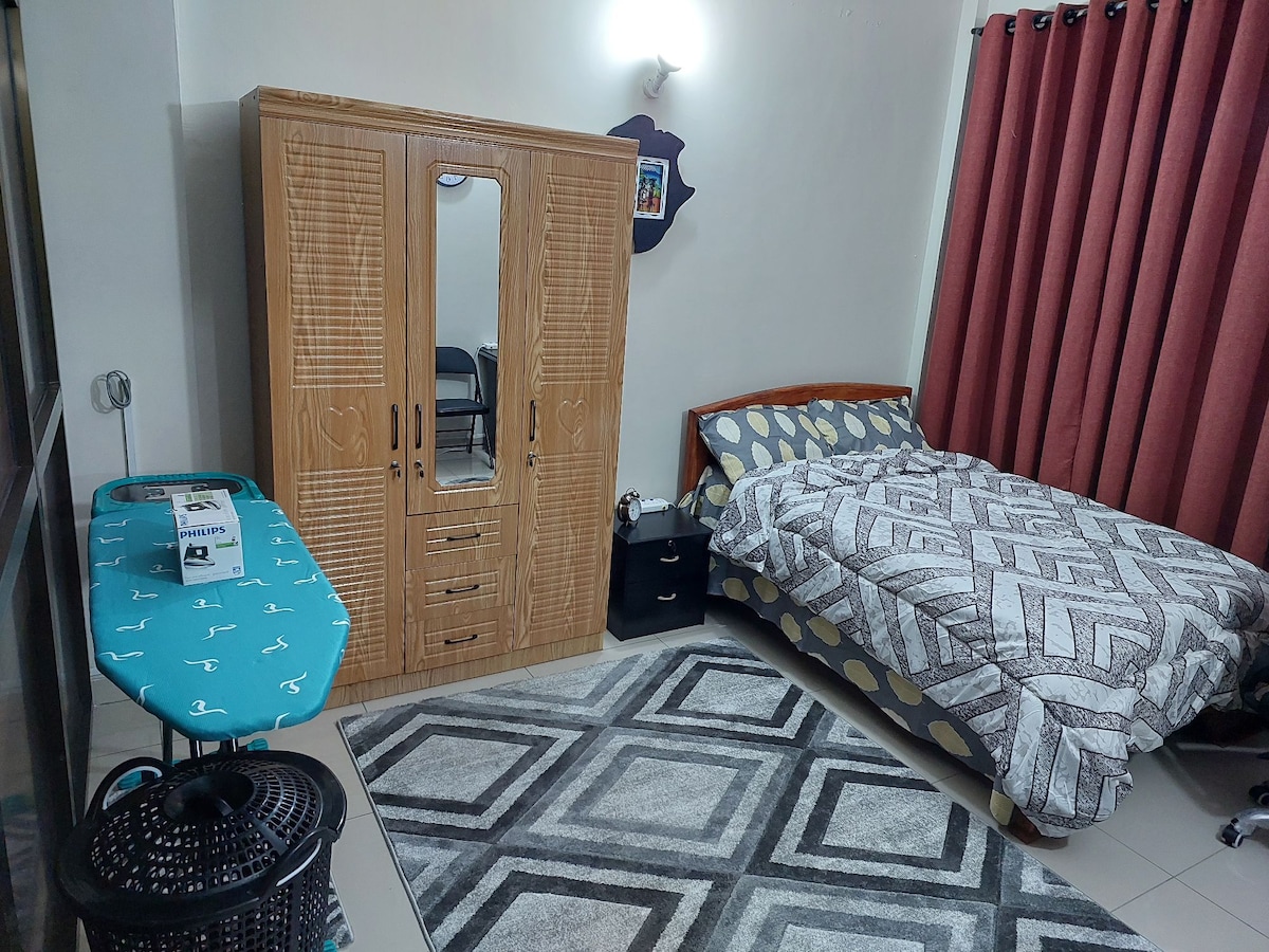 A master bedroom features a spacious bed covered with a patterned blanket, complemented by decorative pillows. A large wardrobe with mirrored doors stands against the wall, while a small nightstand is positioned beside the bed. An ironing table is located nearby, and a geometric rug adds texture to the floor.
