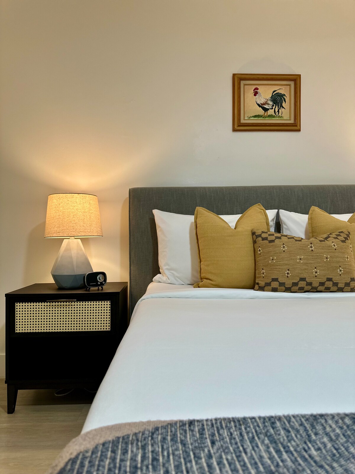 The sleeping area features an upholstered queen bed dressed in crisp white linens, accompanied by decorative pillows in warm tones. A nightstand with a lamp sits beside the bed, casting soft light, while framed artwork adds a touch of color to the wall.
