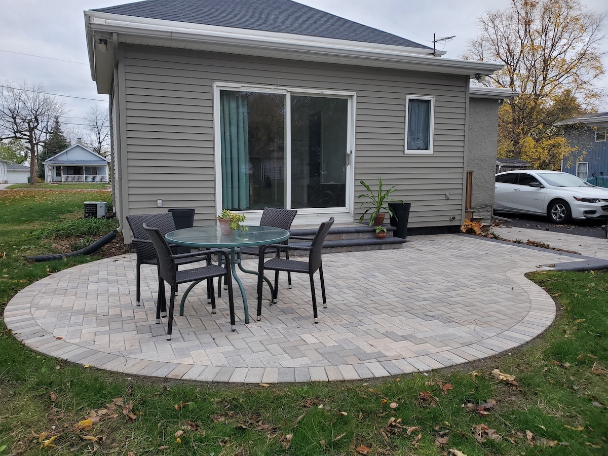 A back patio area is shown, featuring a round glass-top table surrounded by four black chairs. A plant is positioned beside the table. The space is paved with light-colored stones and surrounded by a grassy lawn, providing an inviting outdoor setting.