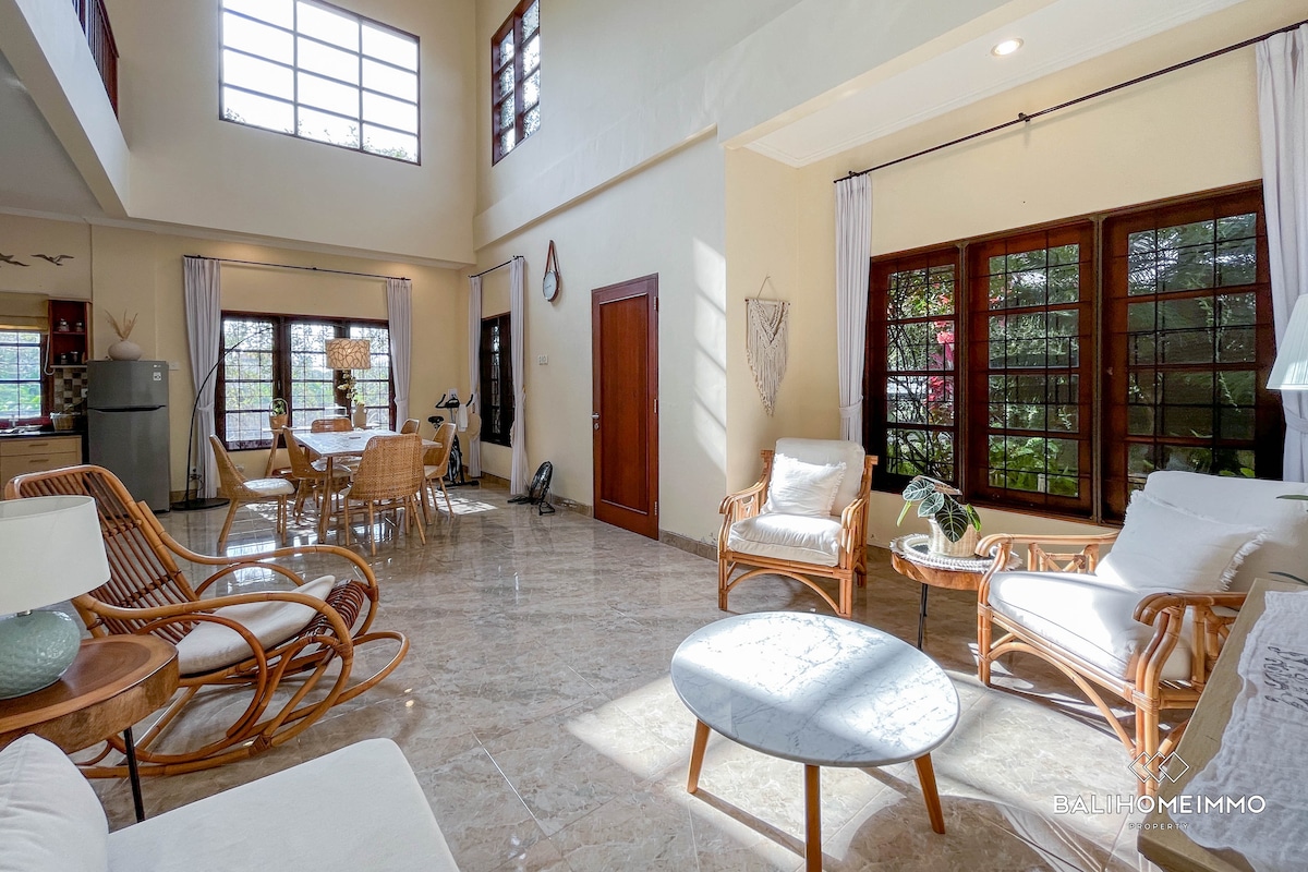 A spacious living room features high ceilings and large windows, allowing natural light to fill the area. Comfortable seating is arranged around a round coffee table. A dining area with a wooden table and woven chairs is visible, creating an inviting space for gatherings.