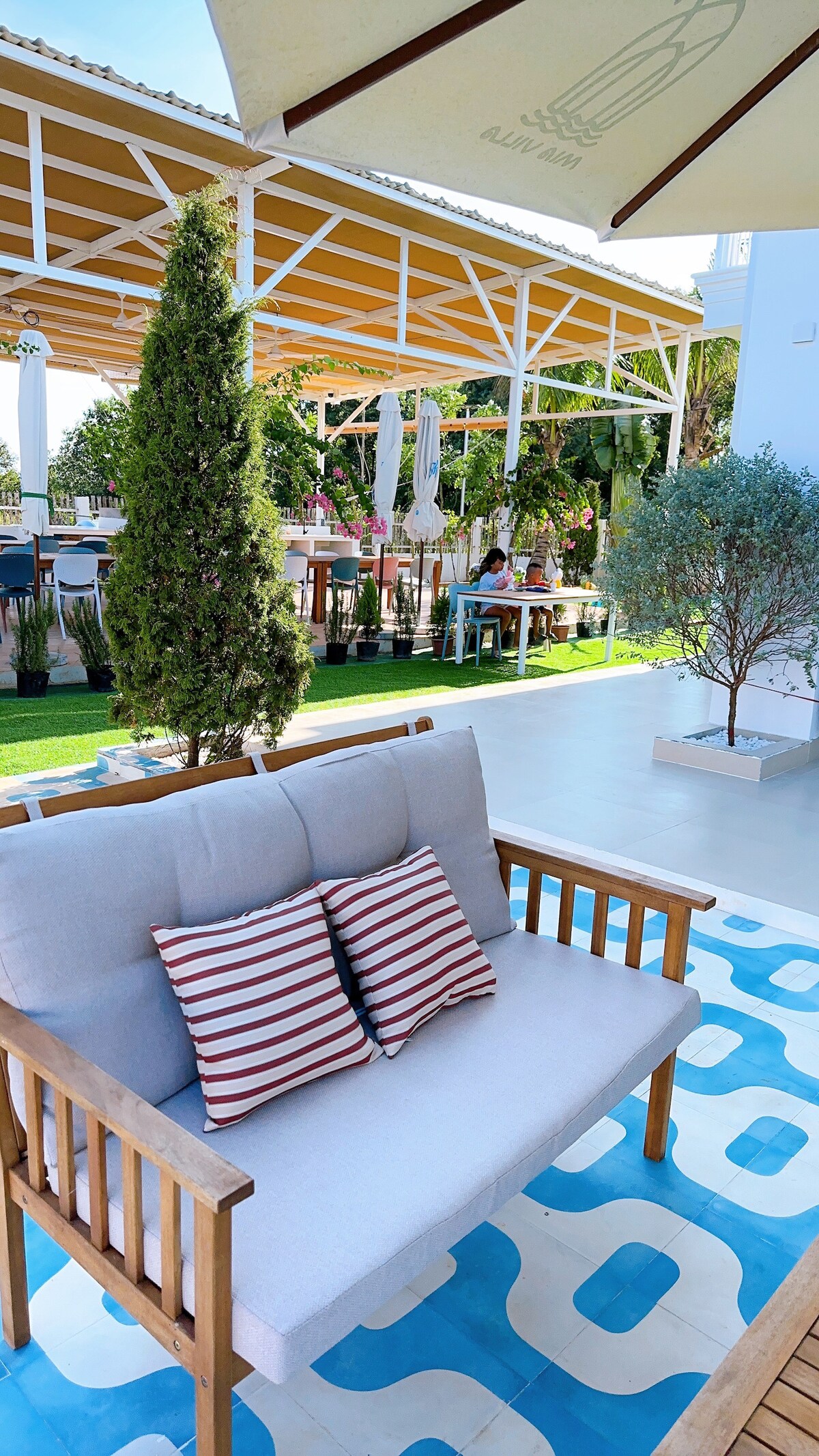 An outdoor seating area features a wooden sofa with striped cushions, positioned on a patterned rug. Lush greenery is present, along with tall trees and umbrellas providing shade. Dining tables are visible in the background, enhancing the inviting outdoor environment.
