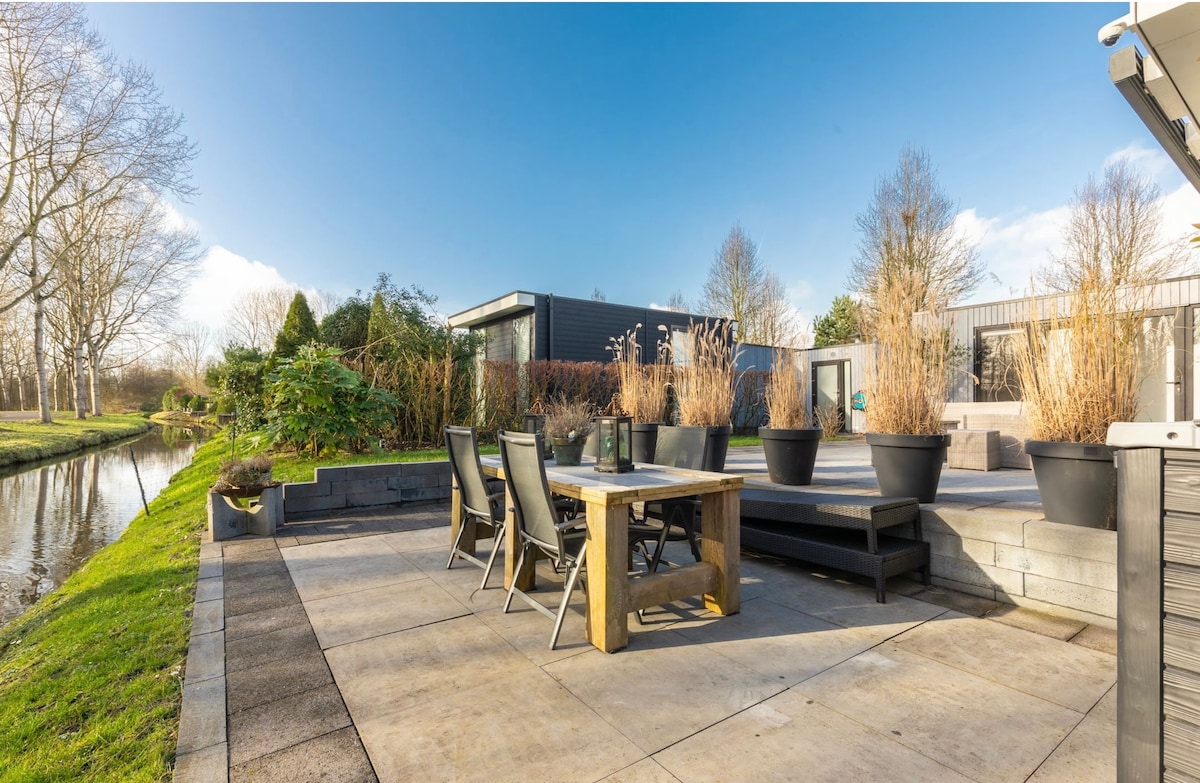 A spacious outdoor terrace features a wooden dining table surrounded by comfortable chairs, with decorative planters adding greenery. The area is bordered by a serene waterway, enhancing the tranquil setting against a clear blue sky.