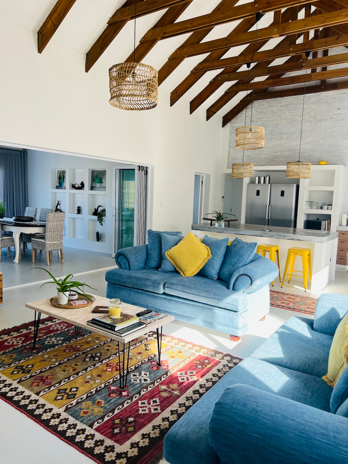 An open-plan living area features high wooden ceilings with exposed beams. A cozy blue couch with a yellow cushion complements a patterned area rug. A coffee table holds decorative items, while a well-equipped kitchen is visible in the background, showcasing sleek appliances and a dining area.