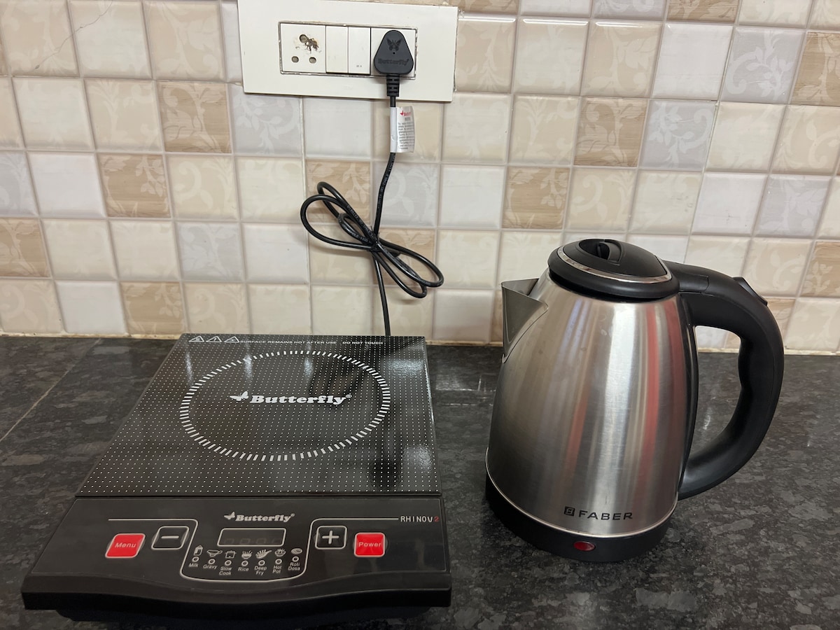 An electric induction cooktop is presented next to a stainless steel kettle on a dark countertop. The cooktop features a digital control panel with clear buttons for easy operation. A power outlet is visible above the countertop, providing convenient access for electrical appliances.