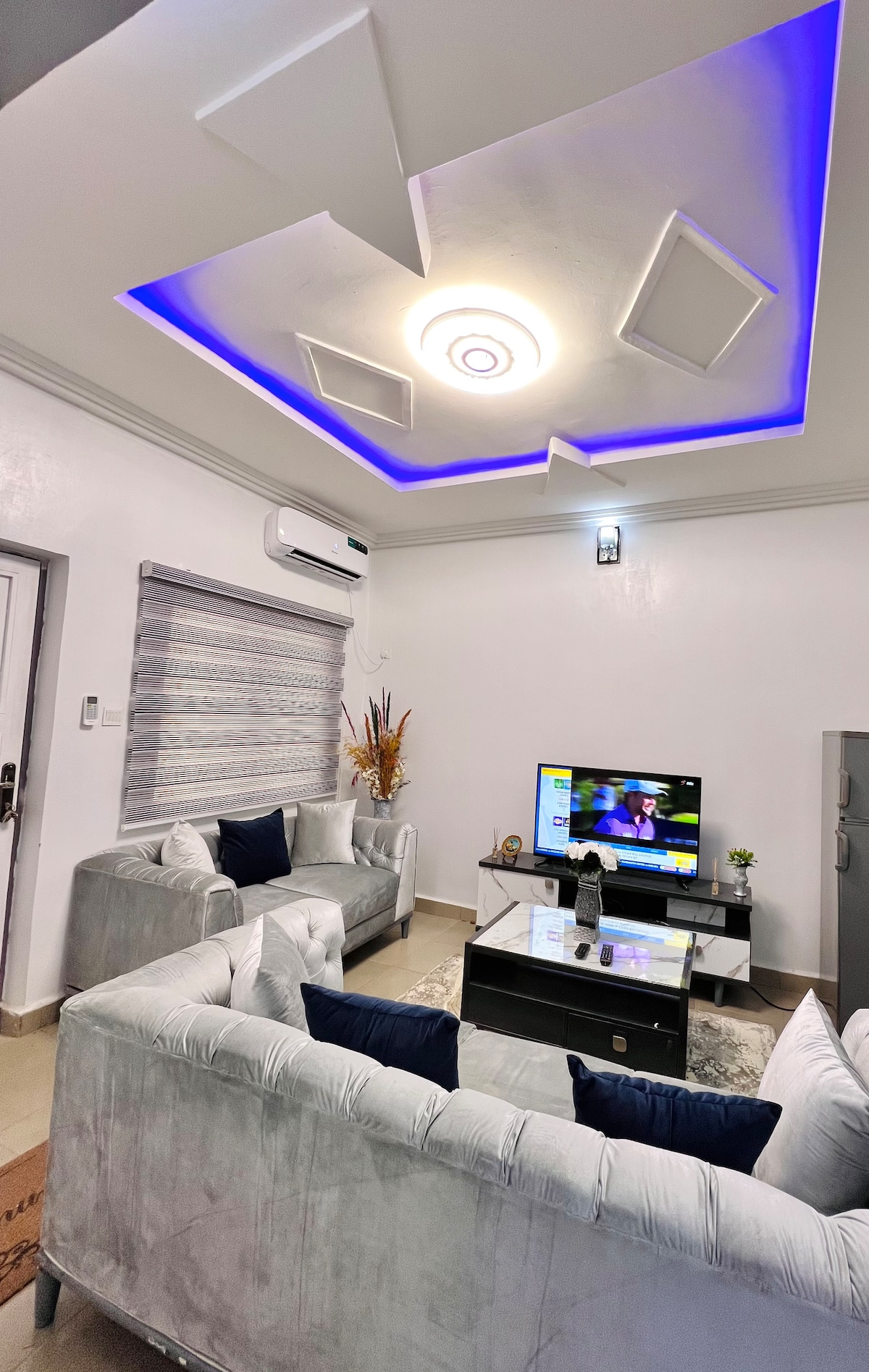 A modern living area features a light-toned sectional sofa paired with deep blue accent pillows. A sleek glass coffee table sits at the center, with a television positioned on a stand against the wall. Soft, ambient lighting enhances the room's inviting atmosphere.