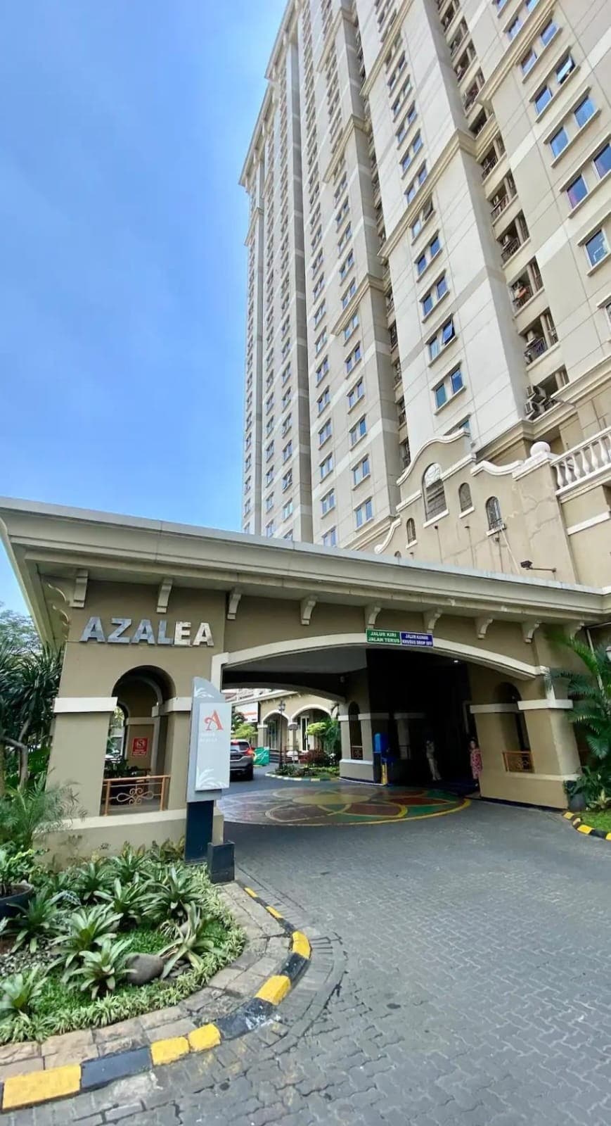 The entrance of the Azalea Tower is showcased, featuring a covered drive-through area with potted plants lining the pathway. The building's architecture is highlighted, displaying a tall structure with a light-colored facade and large windows.