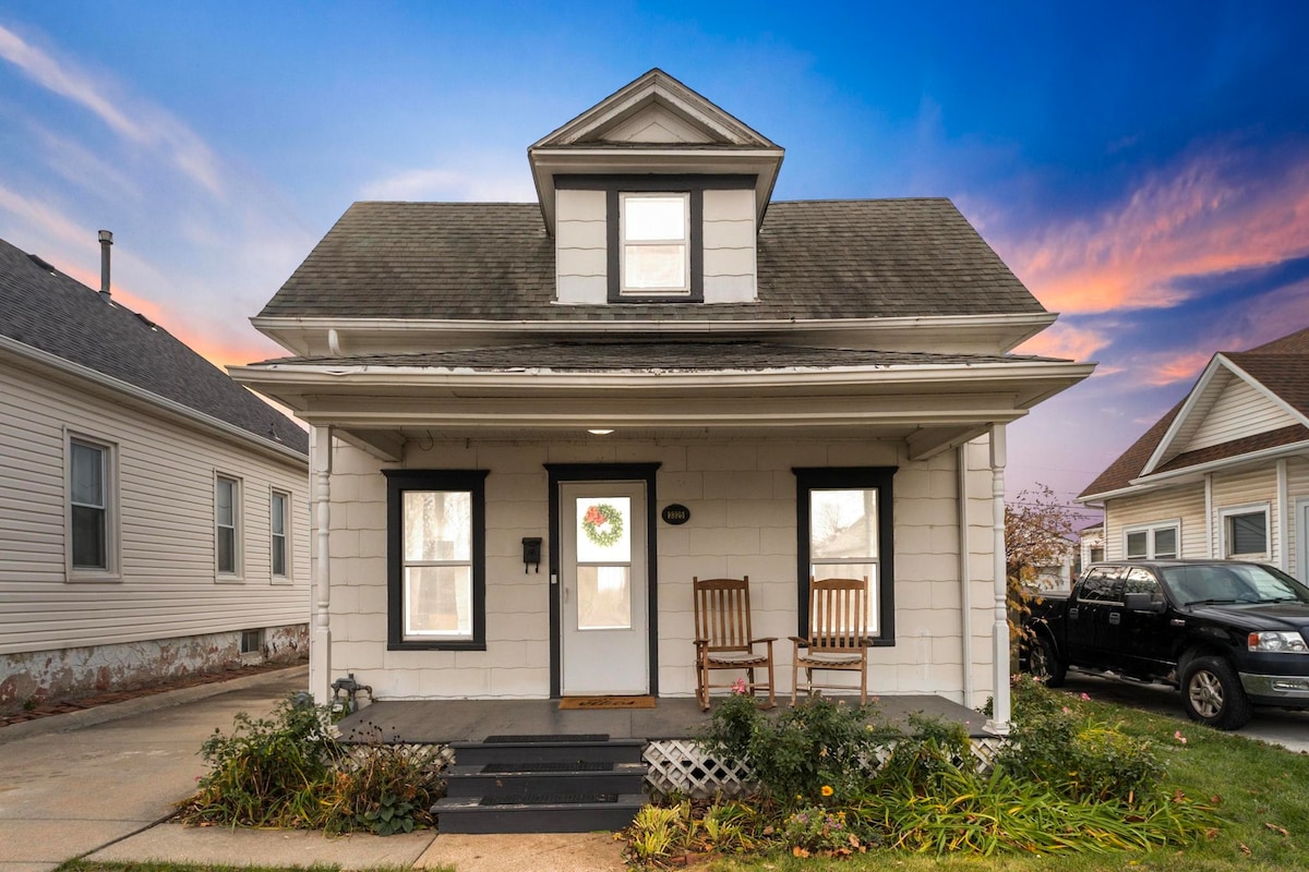 A charming two-story home is presented with a welcoming porch, featuring two rocking chairs. The exterior showcases a light-colored facade with dark trim and large windows, framed by a flower box. The roofline includes a prominent gable, set against a colorful sunset sky.