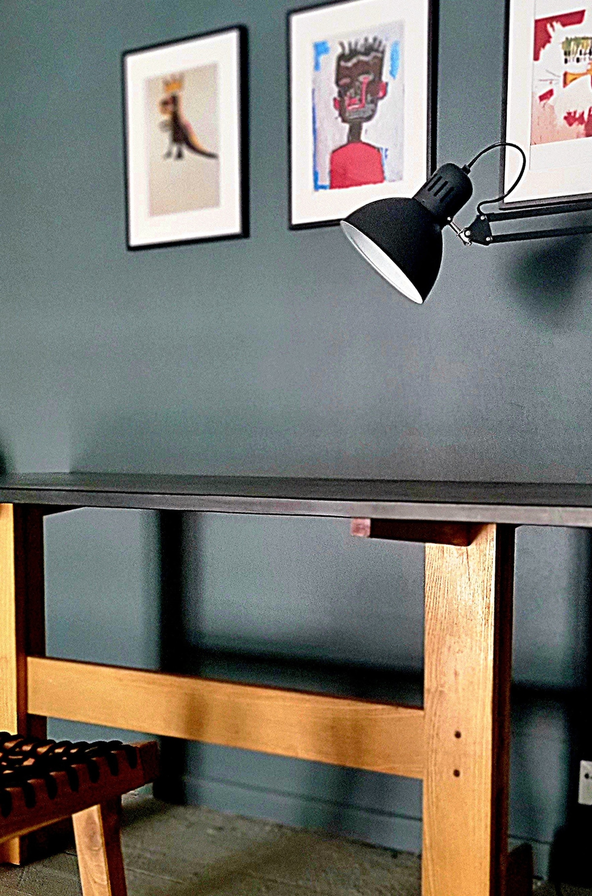 A modern workspace features a sturdy wooden table complemented by a dark table lamp. Abstract artwork adorns the wall above, adding a touch of creativity to the space. The contrasting dark wall enhances the contemporary design.