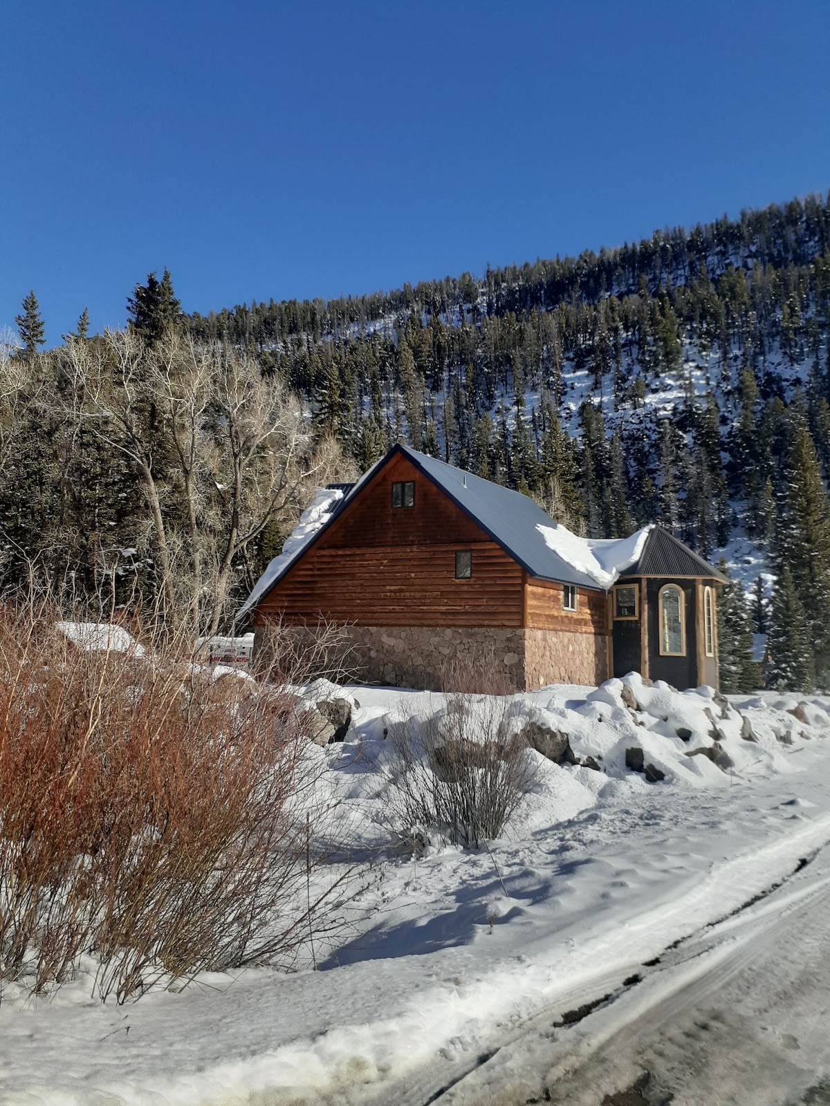 A rustic cabin is nestled among snow-covered terrain, framed by tall evergreen trees. The building features a combination of wooden and stone siding, with a sloped roof adorned with fresh snow. Clear blue skies provide a bright backdrop to the winter landscape.