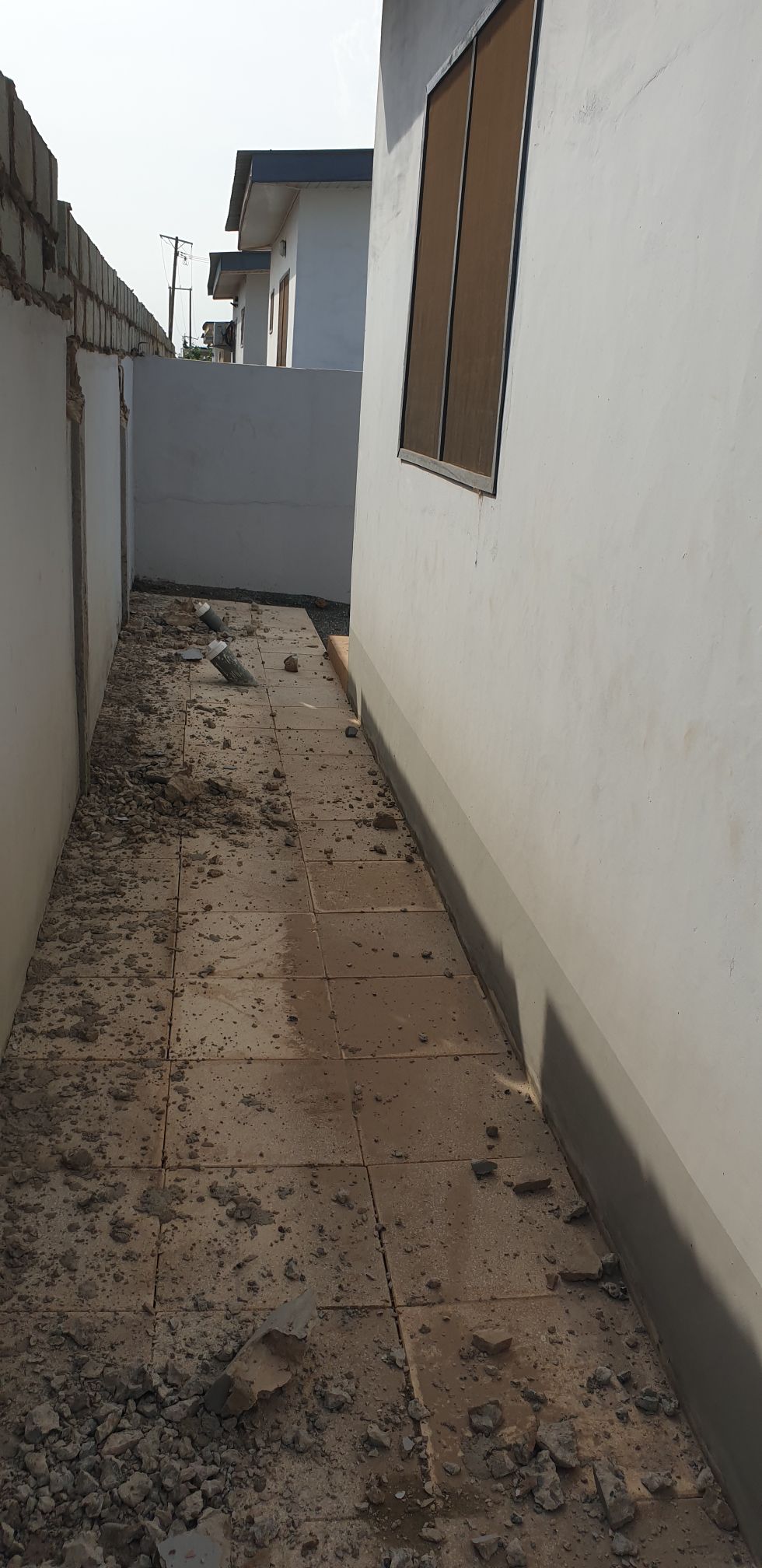 A narrow pathway is flanked by plain white walls, with a window allowing light to enter the space. The ground is paved with tiles, which are slightly uneven and scattered with debris, creating a utilitarian environment.
