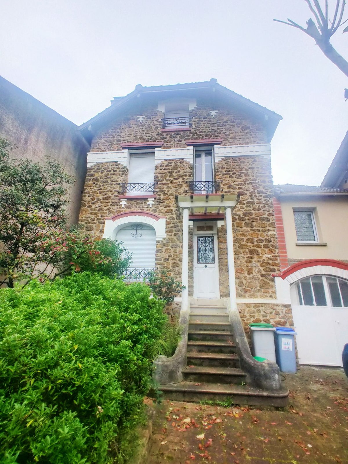 The exterior of the villa showcases a charming stone facade, highlighted by decorative balcony railings. A staircase leads to the main entrance, framed by lush greenery on either side. The surrounding area features additional residential buildings, contributing to a serene neighborhood atmosphere.