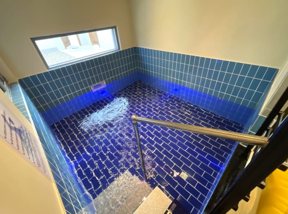 An indoor heated pool is enclosed by vibrant blue tiles. Clear water reflects soft lighting, creating a calming ambiance. A stainless steel railing provides safety access to the pool, while natural light filters through the window, enhancing the serene atmosphere.