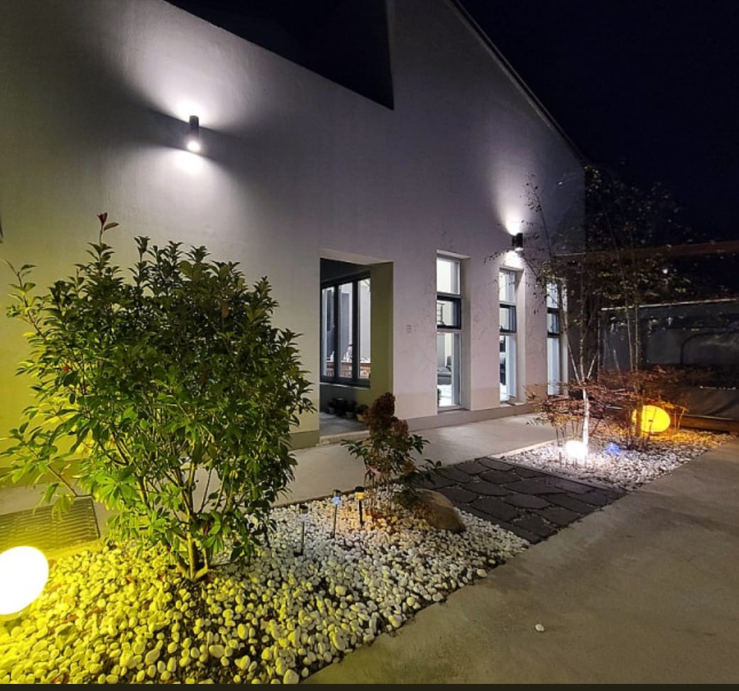 An outdoor area is visibly illuminated at night, featuring neatly arranged stones and a landscaped garden. Soft lighting enhances the pathways and plants, creating a serene atmosphere. Multiple windows suggest nearby indoor spaces, contributing to the overall ambiance.